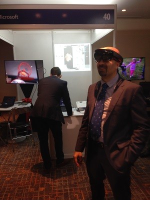 Ông Rafic, đeo cả kính cận và Hololen, đang nhìn thấy một con chuột đang chạy trong bánh xe quay màu đỏ vốn hoàn toàn vô hình nếu nhìn bằng mắt thường trong không gian thực của khách sạn Sheraton (hình ảnh này được hiển thị ra màn hình phía sau bên phải của ông) Ông Rafic, đeo cả kính cận và Hololen, đang nhìn thấy một con chuột đang chạy trong bánh xe quay màu đỏ vốn hoàn toàn vô hình nếu nhìn bằng mắt thường trong không gian thực của khách sạn Sheraton (hình ảnh này được hiển thị ra màn hình phía sau bên phải của ông)